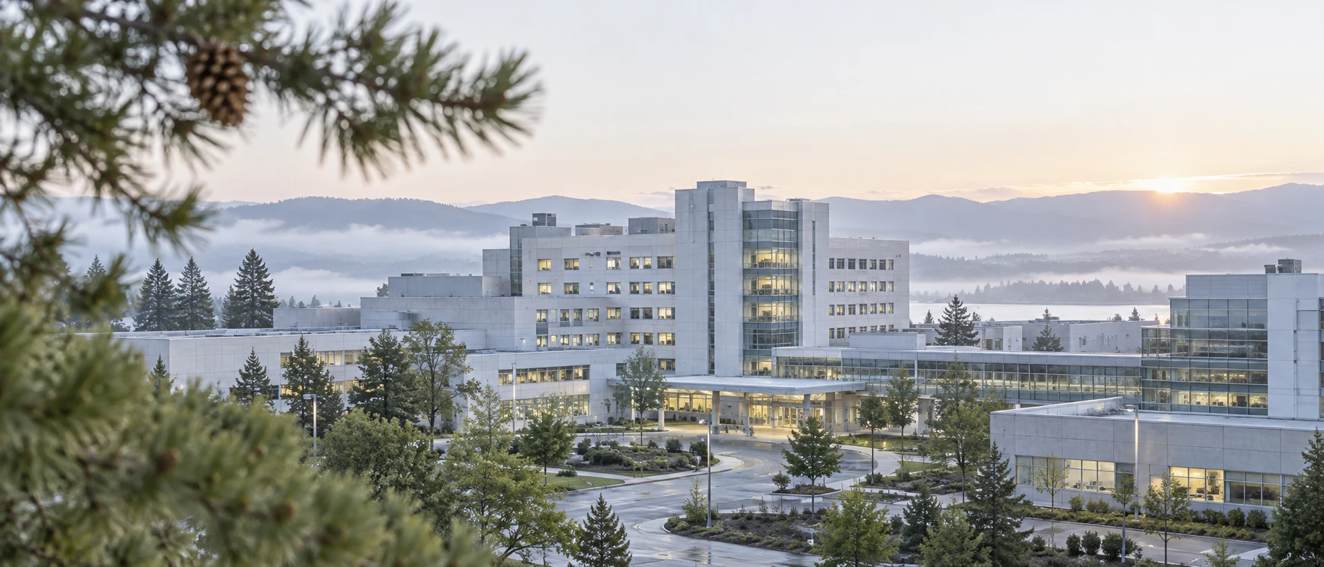 Bright dawn over a modern teaching-hospital campus seen from a slight elevation: pale sky, morning haze, a few windows still lit from the ending night shift. In the near foreground,…
