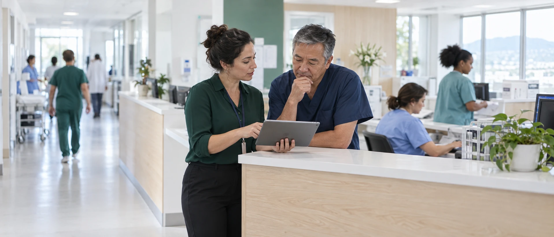 Two colleagues talk over a tablet at a nurses' station in a bright hospital while staff walk past.