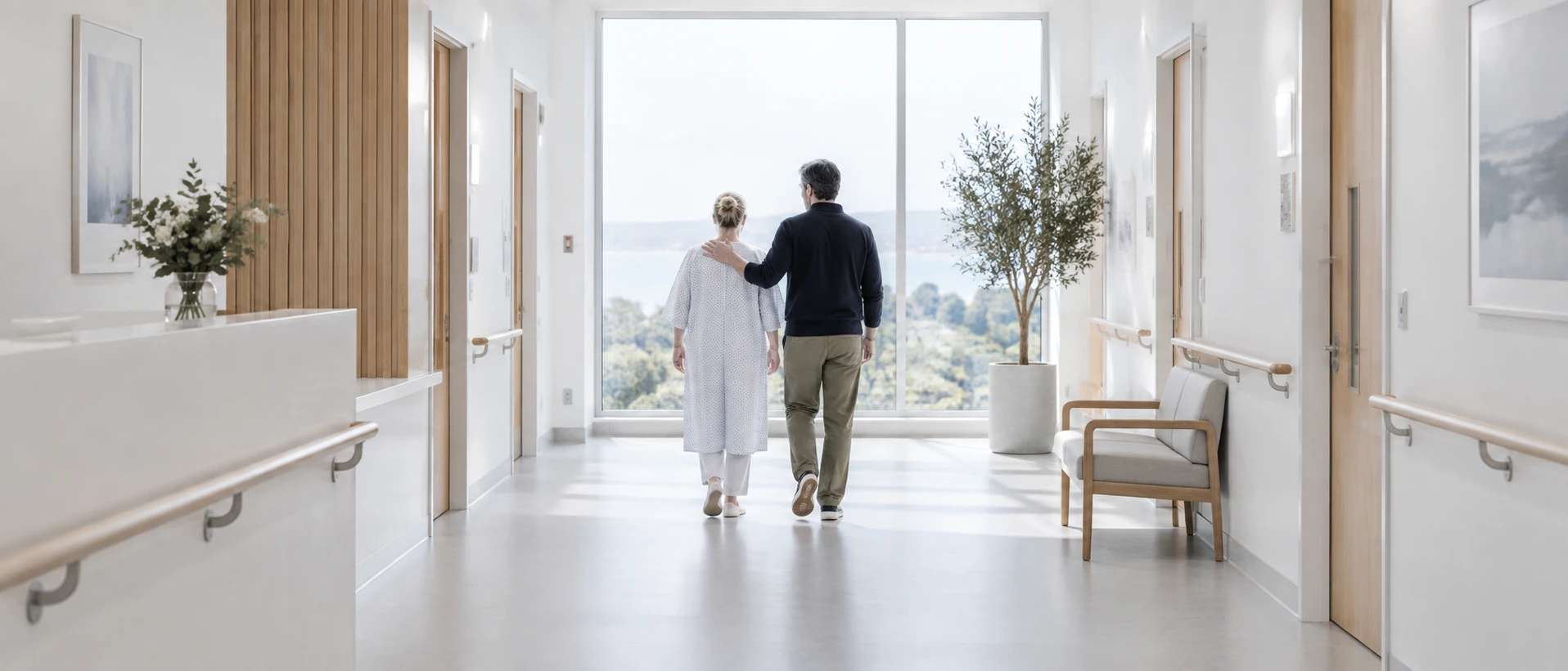 A patient and a close companion walk away from the camera down a bright hospital corridor towards a tall window, the companion's hand lightly on her shoulder. Ultra-wide frame, soft…