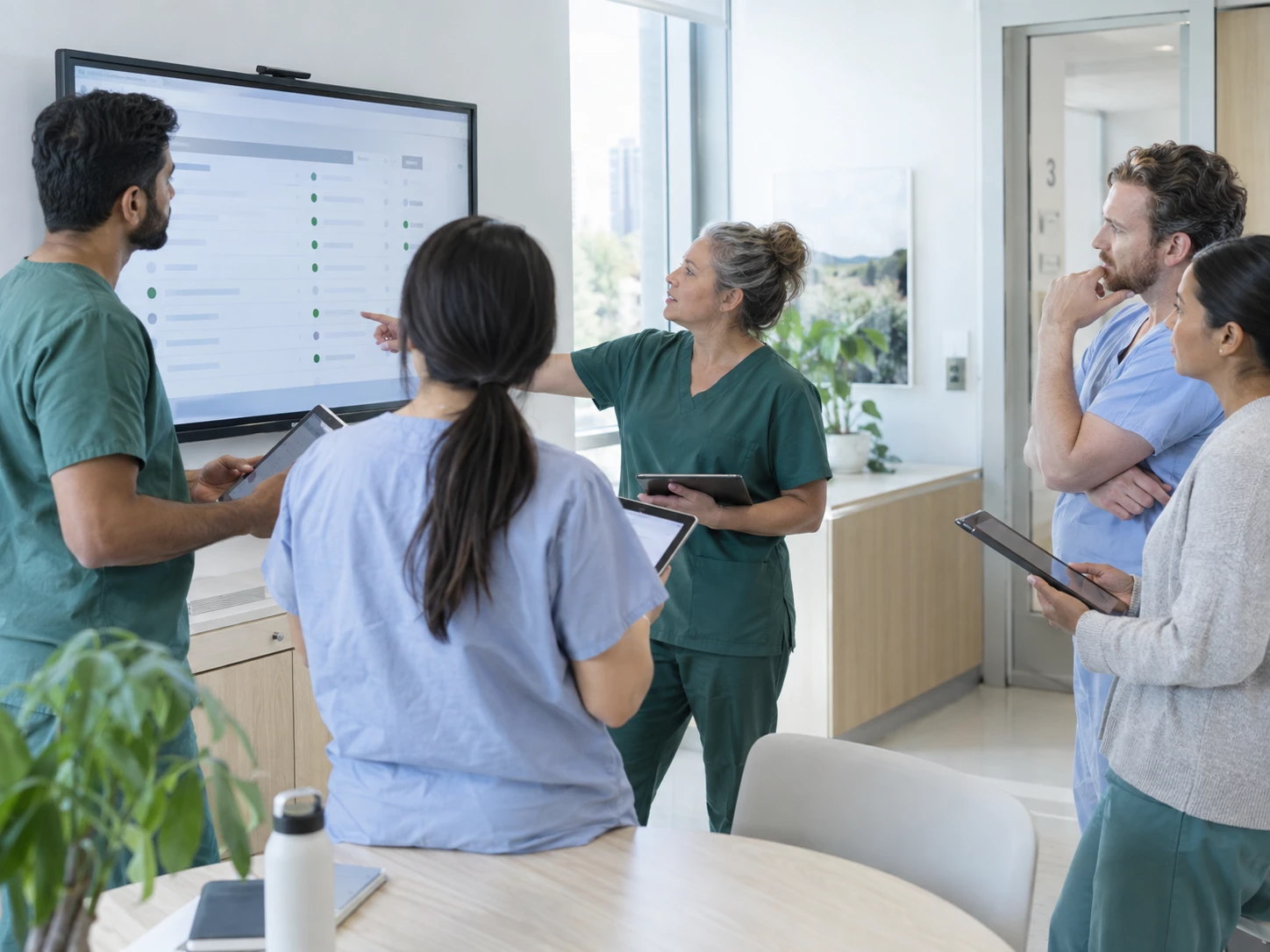 A department briefing in a bright teaching clinic: a small team stands around a wall display showing an abstract list of cases, some rows marked with a pine-green indicator. Tablets in…