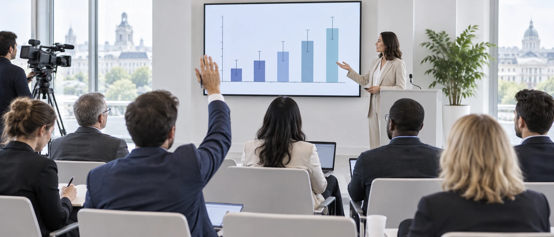 A briefing for journalists and analysts in a bright room: a presenter beside a screen with an abstract chart showing bars with confidence-interval whiskers, a raised hand in the audience.…